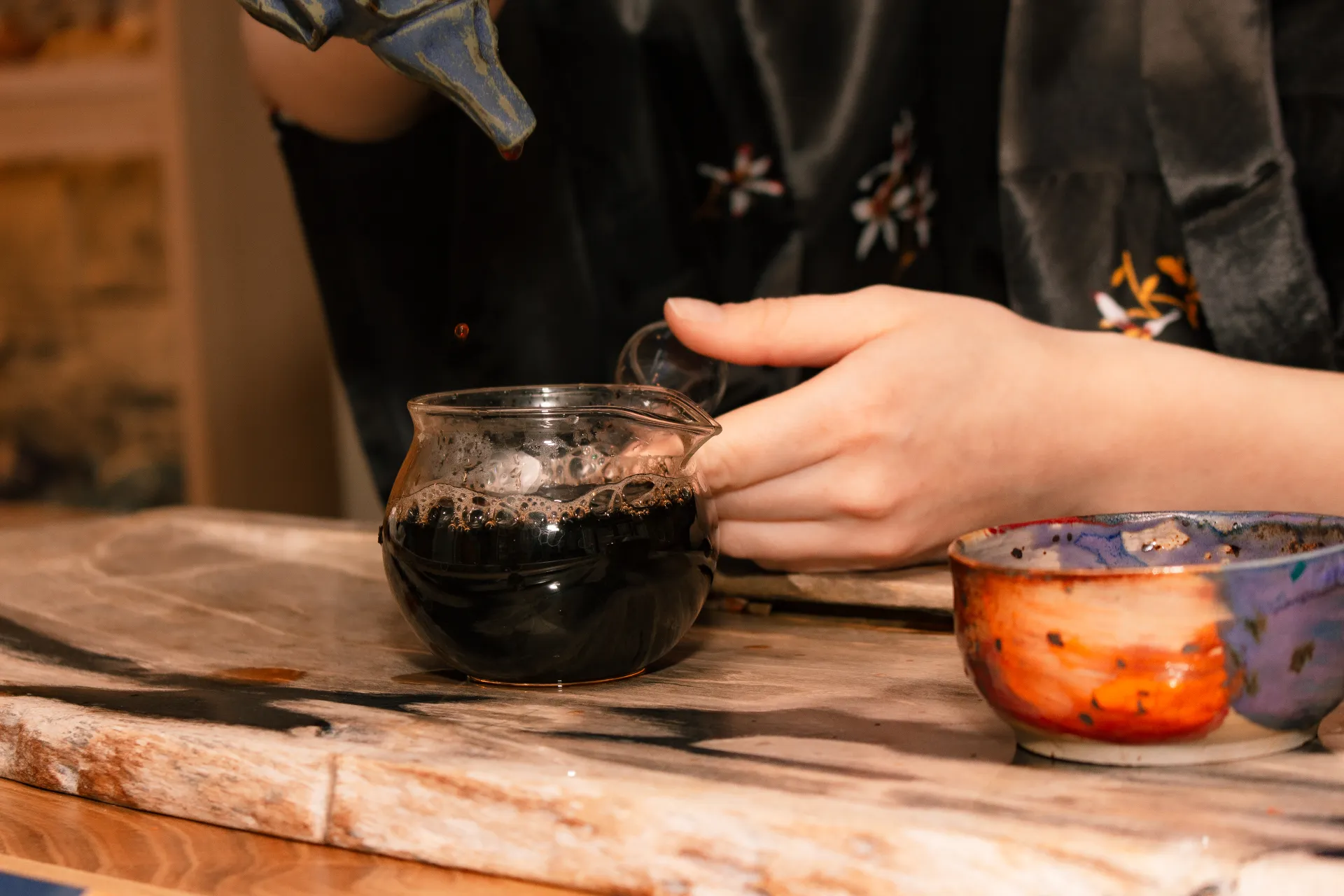 Primer plano de una persona vertiendo té negro preparado de una tetera en una taza de vidrio sobre una superficie de madera, con un cuenco de cerámica colorido cerca.