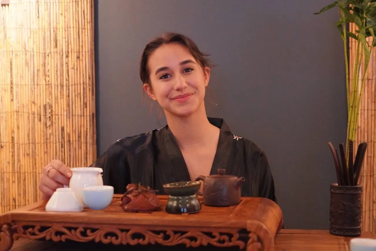 Young woman with dark hair smiling at a tea ceremony, surrounded by traditional teaware on a carved wooden tray, with bamboo decor in the background.