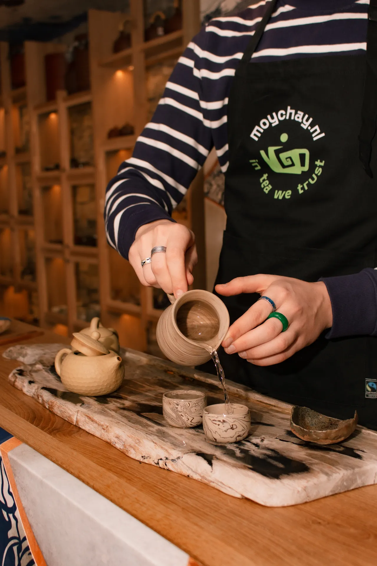 Person pouring hot water into a ceramic tea cup on a wooden table, wearing a black apron with 'Moychay.nl' logo and striped shirt, in a cozy tea shop.
