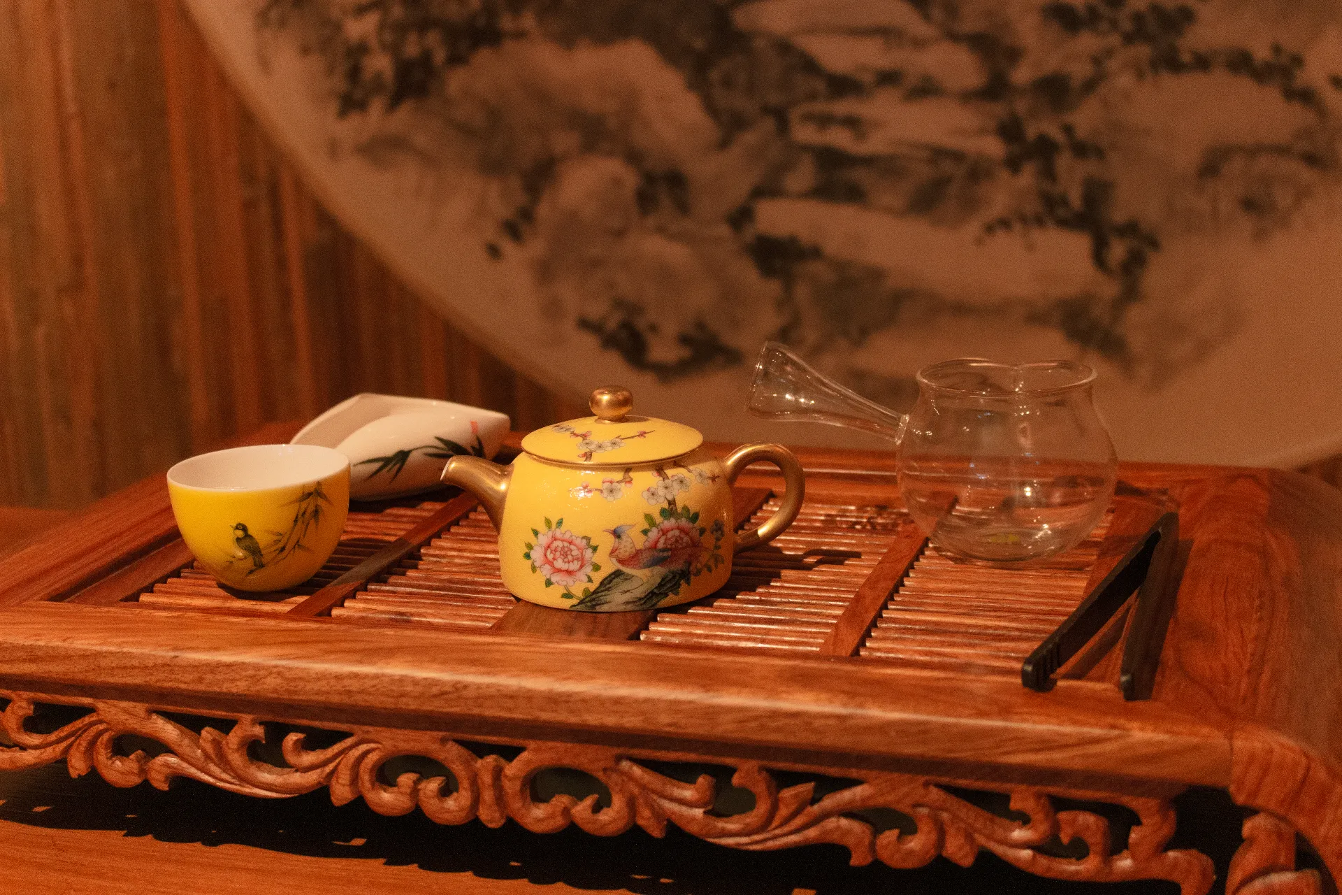 Elegante juego de utensilios chinos para té con una tetera amarilla, taza a juego, un pequeño recipiente con tapa, una jarra de vidrio y pinzas sobre una bandeja de madera.