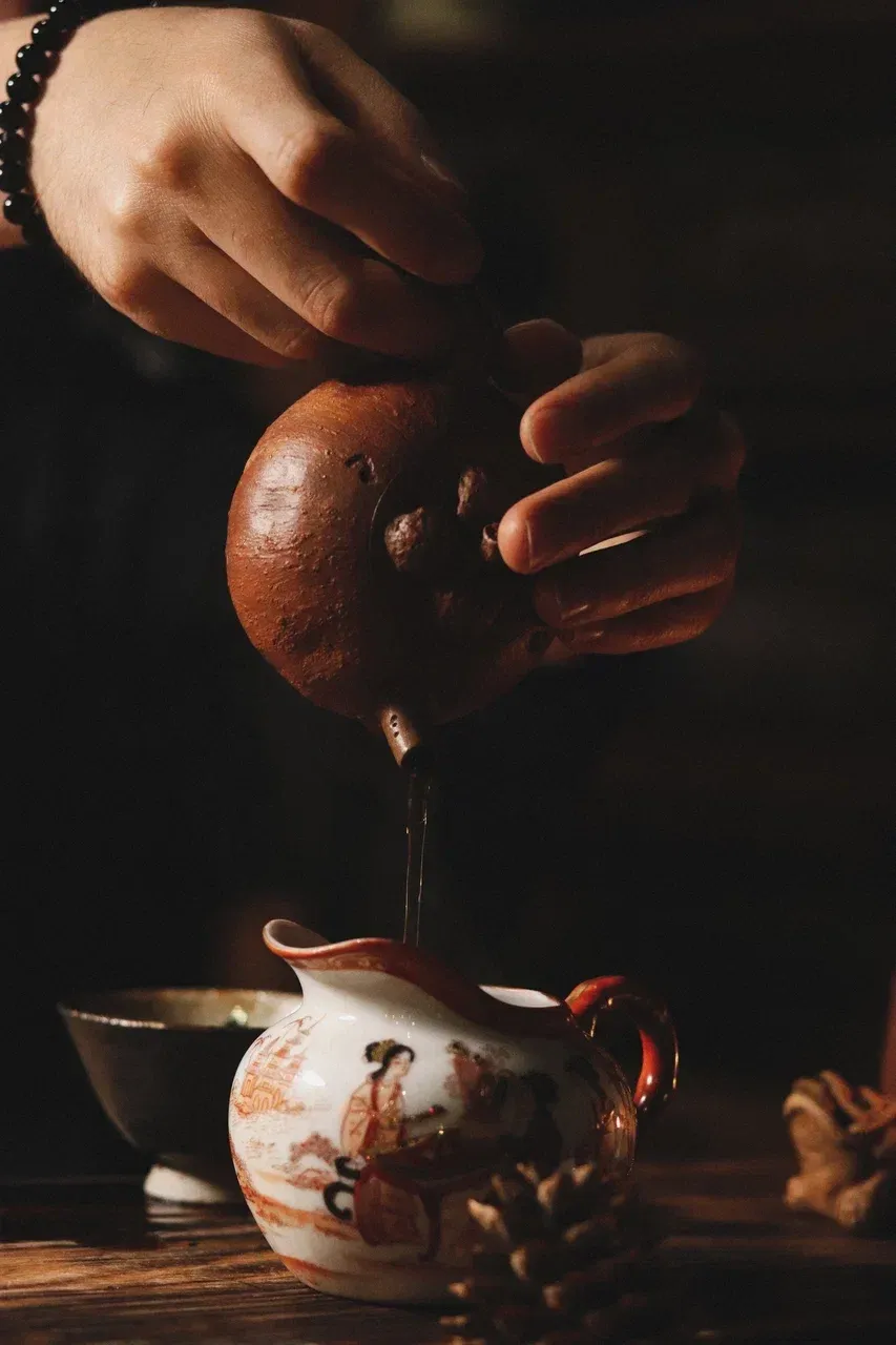Hands pour dark tea from a traditional Gaiwan into a porcelain teapot depicting historic Asian figures, emphasizing mindfulness and ritual.